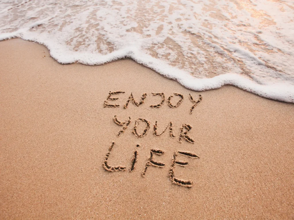 In den Sand geschriebener Satz „Enjoy your life“ wird vom Meer erreicht – Symbol für Vergänglichkeit und Gegenwärtigkeit.