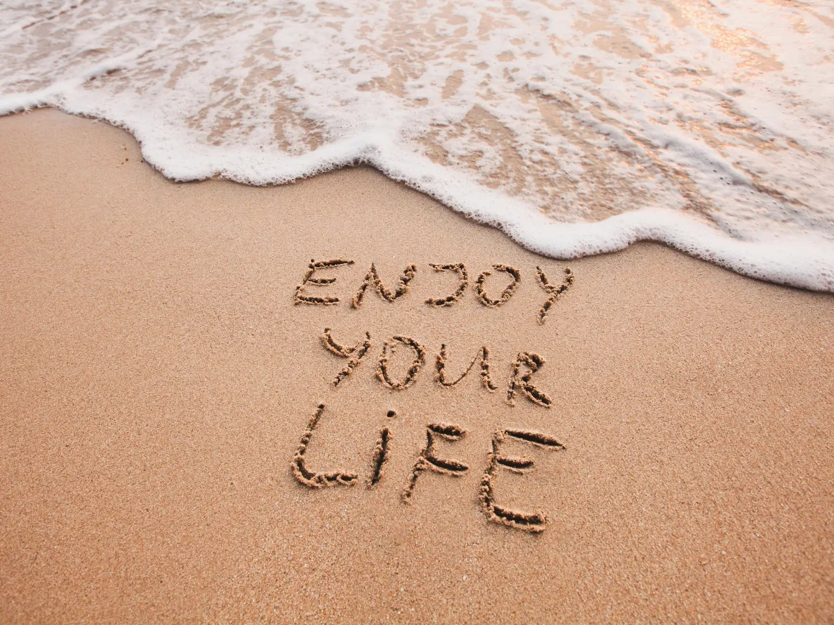 In den Sand geschriebener Satz „Enjoy your life“ wird vom Meer erreicht – Symbol für Vergänglichkeit und Gegenwärtigkeit.