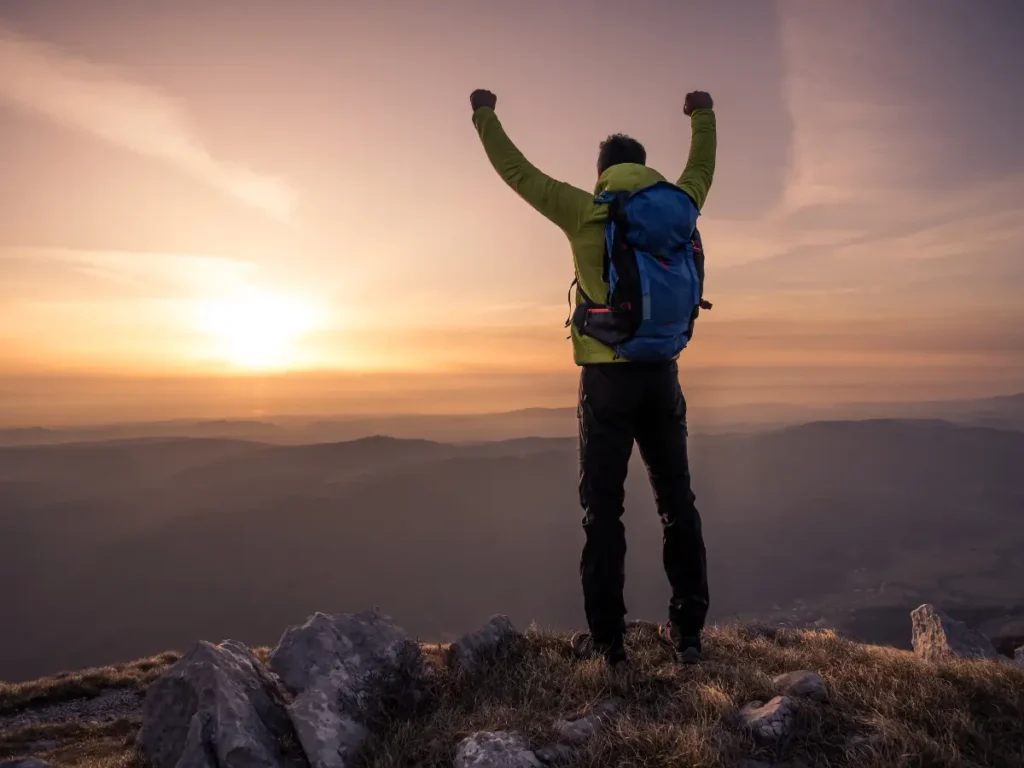 Ein Wanderer mit erhobenen Armen auf einem Berggipfel bei Sonnenaufgang – Symbol für Kraft, innere Stärke, Zielerreichung und Entschlossenheit